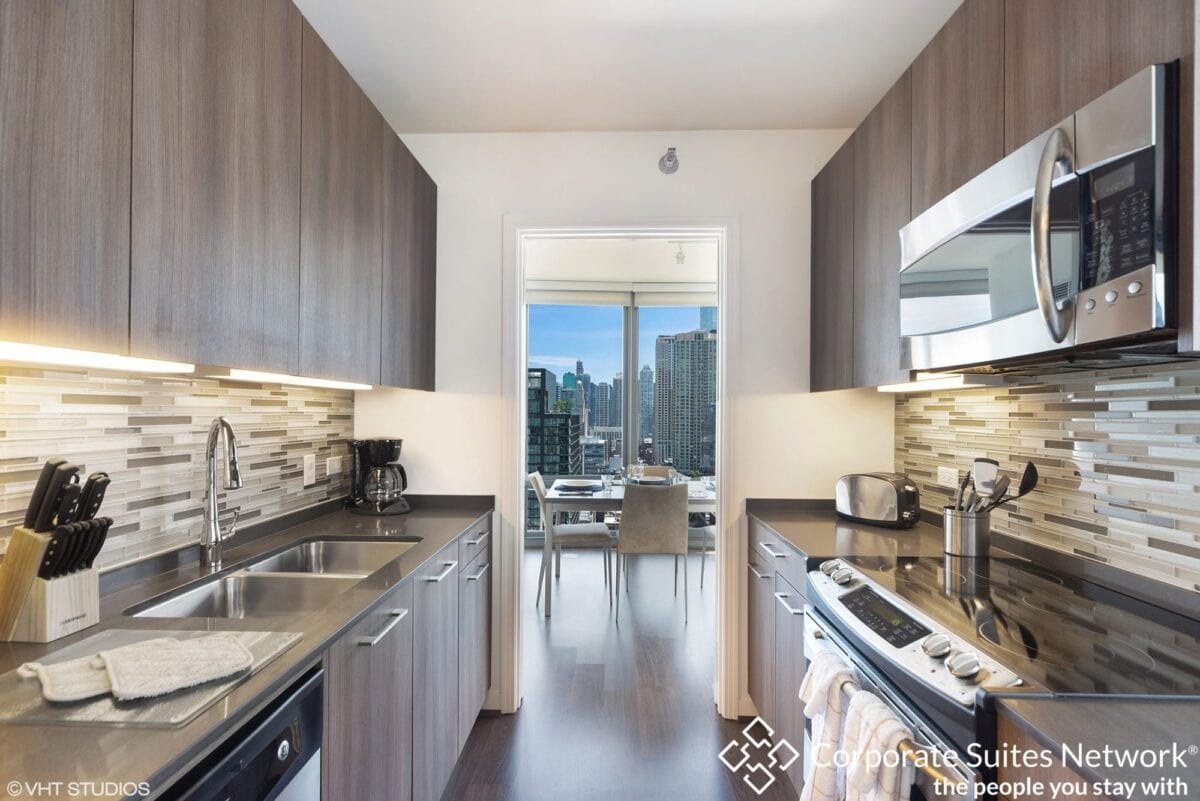 Modern galley kitchen with wood cabinets, stainless steel appliances, and tile backsplash; dining area with city view visible through doorway.