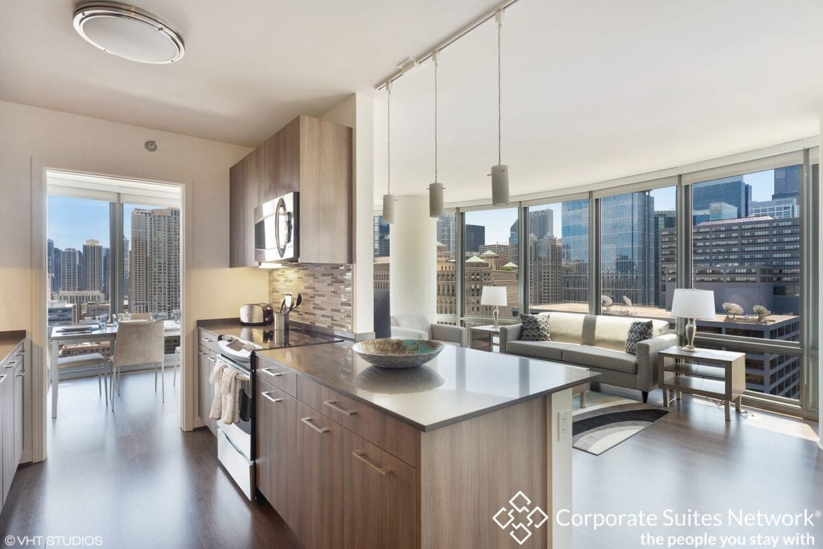 Modern apartment kitchen with stainless steel appliances and island, open to a bright living area with large windows and city skyline views.