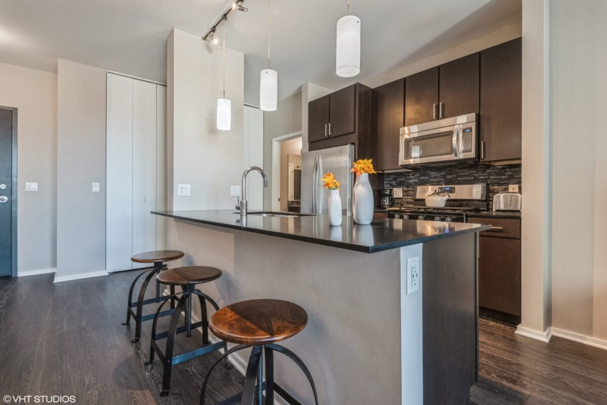 Modern kitchen with dark cabinets, stainless steel appliances, a black island with sink, three wooden stools, and pendant lighting. Two vases with orange flowers are on the countertop.
