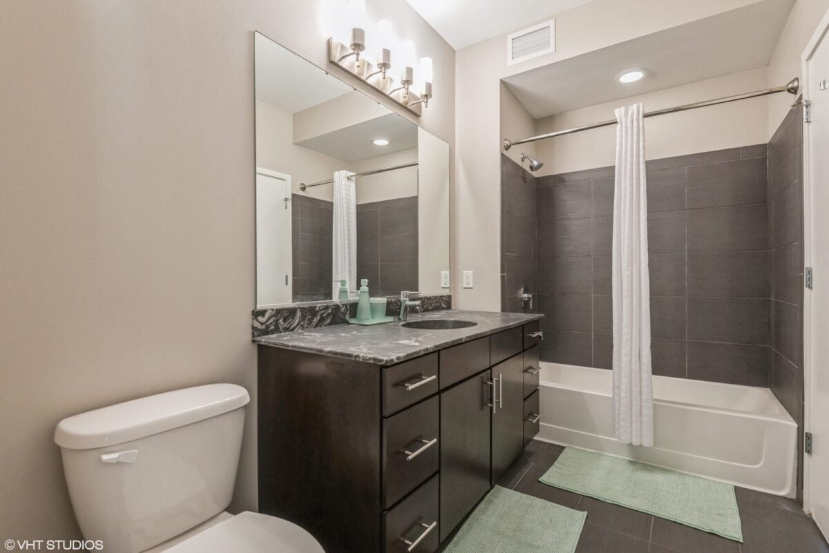 Modern bathroom with a dark wood vanity, white countertop, large mirror, toilet, bathtub with gray tile surround, and white shower curtain. Two green bath mats on the floor.