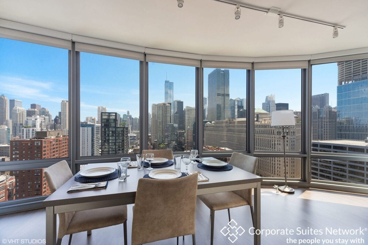 Dining table set for four in a modern apartment with floor-to-ceiling windows overlooking a city skyline on a clear day.