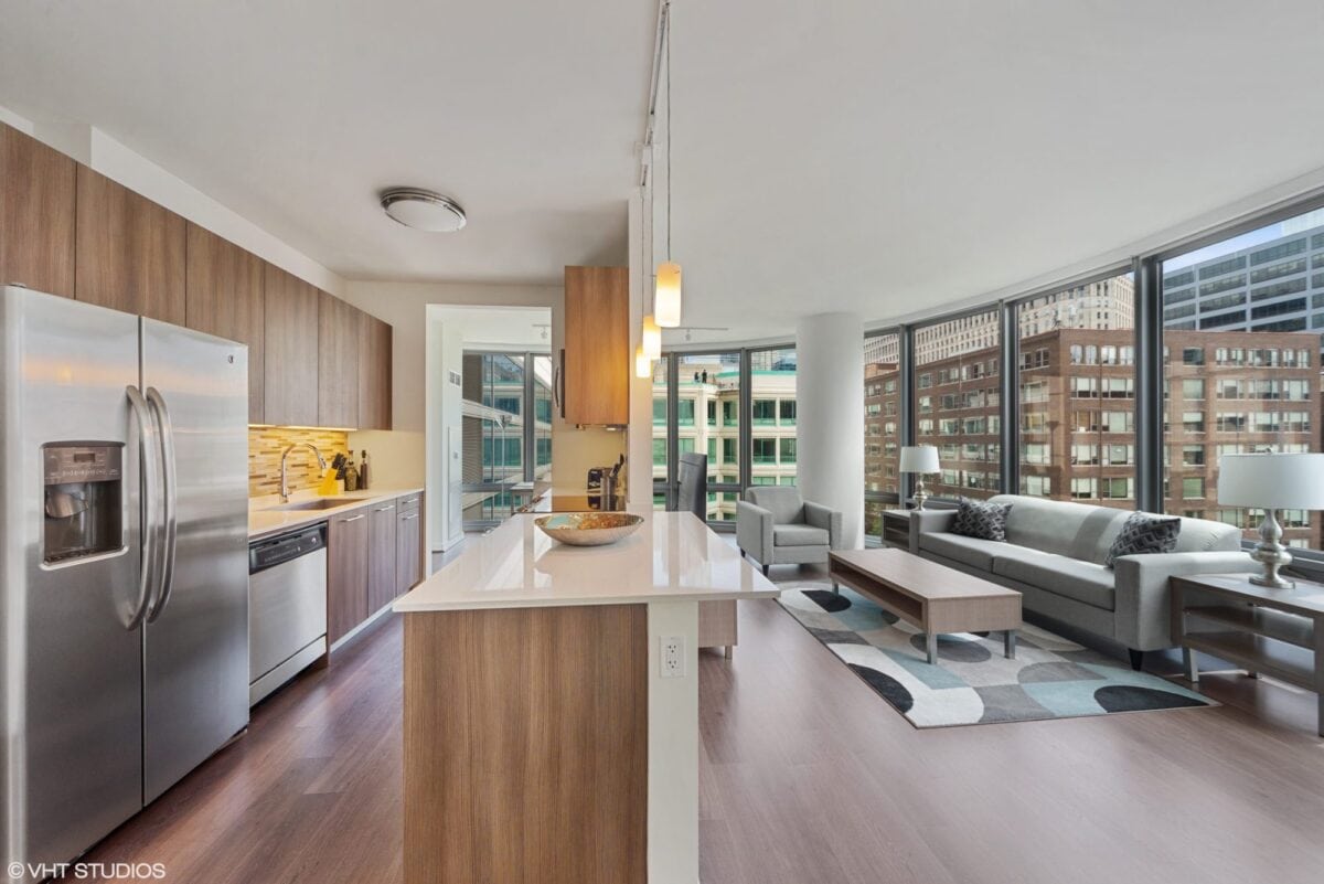 Modern open-concept kitchen and living room with wood cabinetry, stainless steel appliances, large windows, and city views. Neutral décor with contemporary furniture and natural light.
