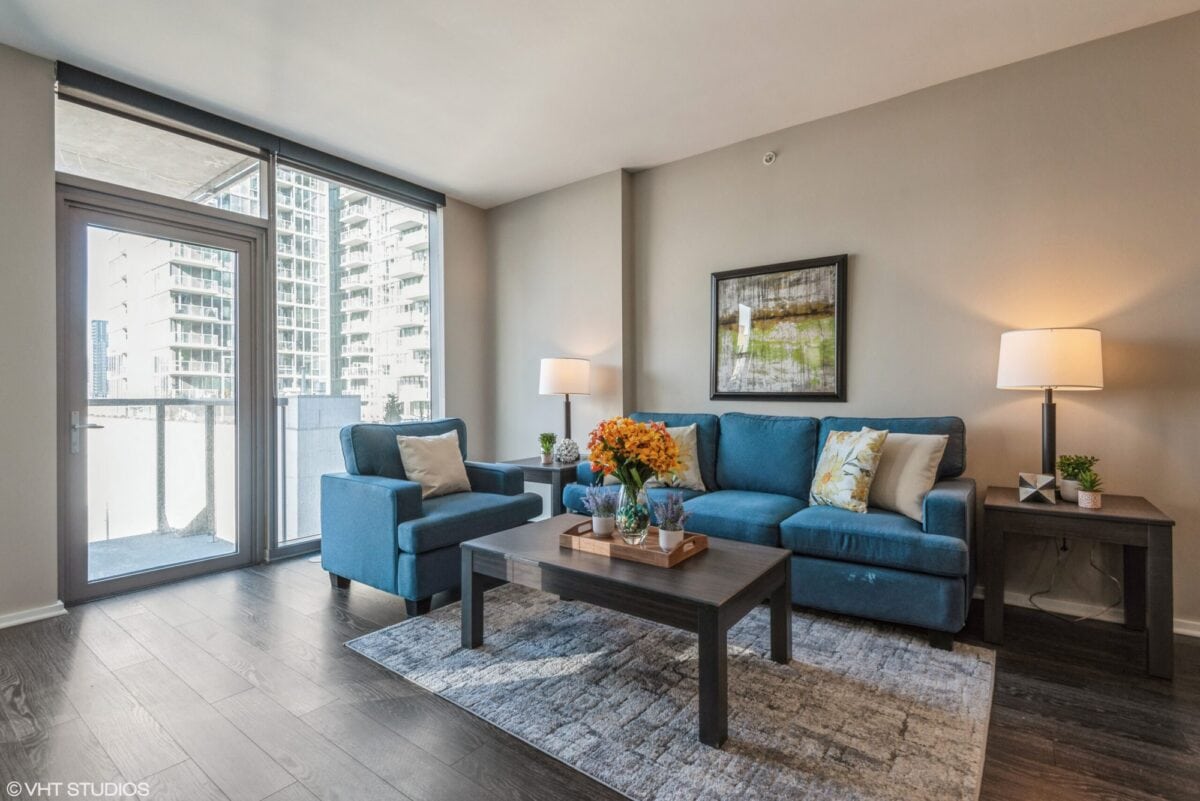 Modern living room with blue sofa and armchair, coffee table with flowers, two lamps, framed art, large windows, and balcony view of city buildings.