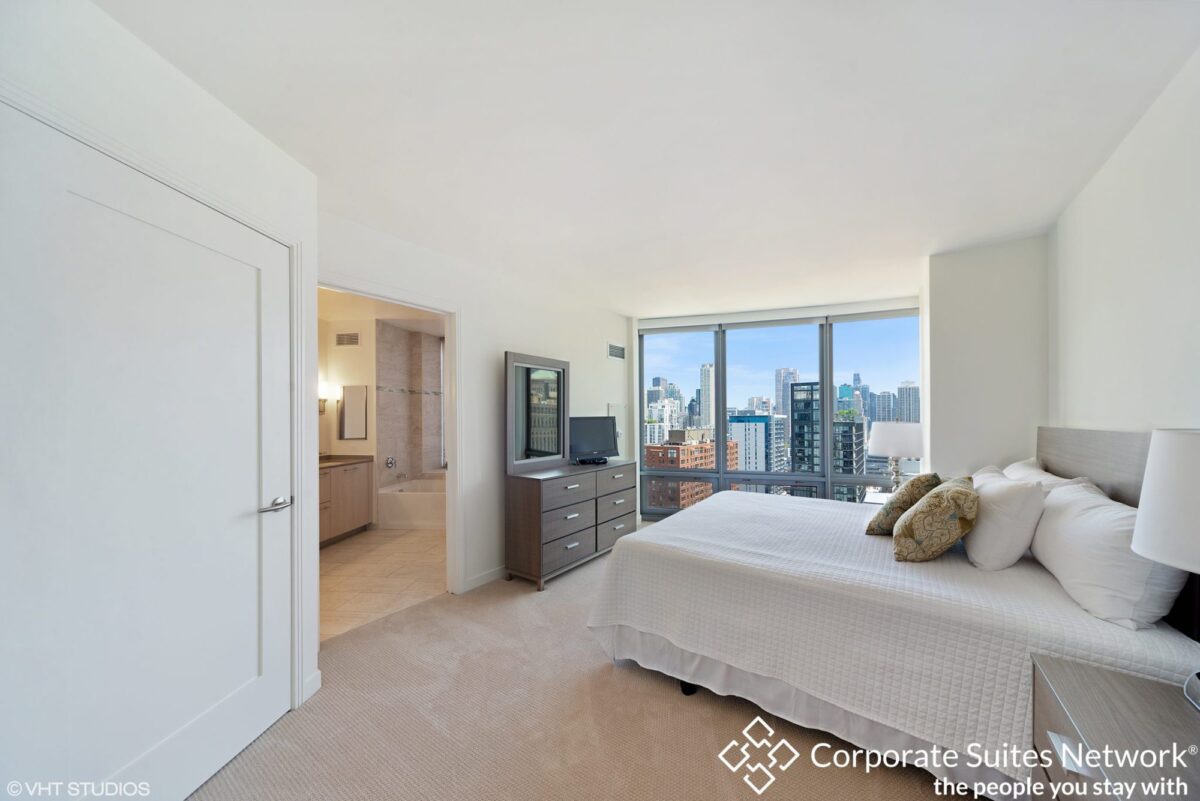 Modern bedroom with large windows, city skyline view, neutral decor, dresser with TV, and attached bathroom; Corporate Suites Network logo in the corner.