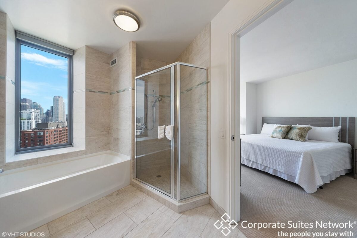 Modern bathroom with a glass shower, bathtub, and city view window; adjacent bedroom with a large bed and neutral decor visible through open door.