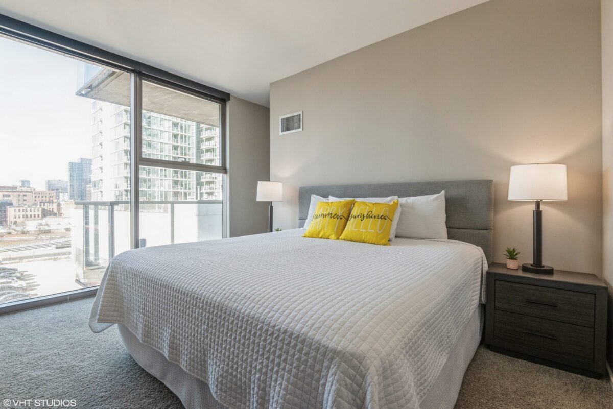 Modern bedroom with large window, king-size bed with white bedding, two bedside tables with lamps, and cityscape view outside. Yellow pillows add a pop of color.
