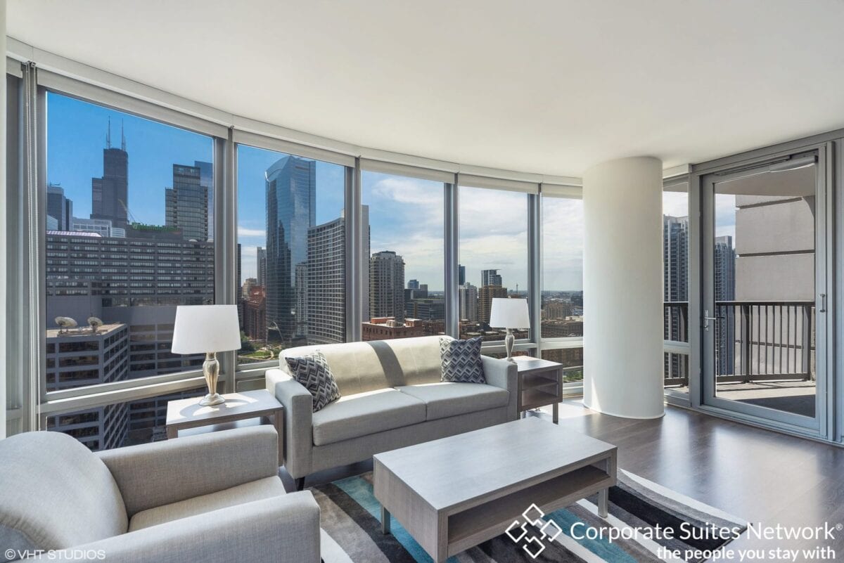 Modern living room with large windows, city skyline view, neutral-toned furniture, a coffee table, and an adjacent balcony.