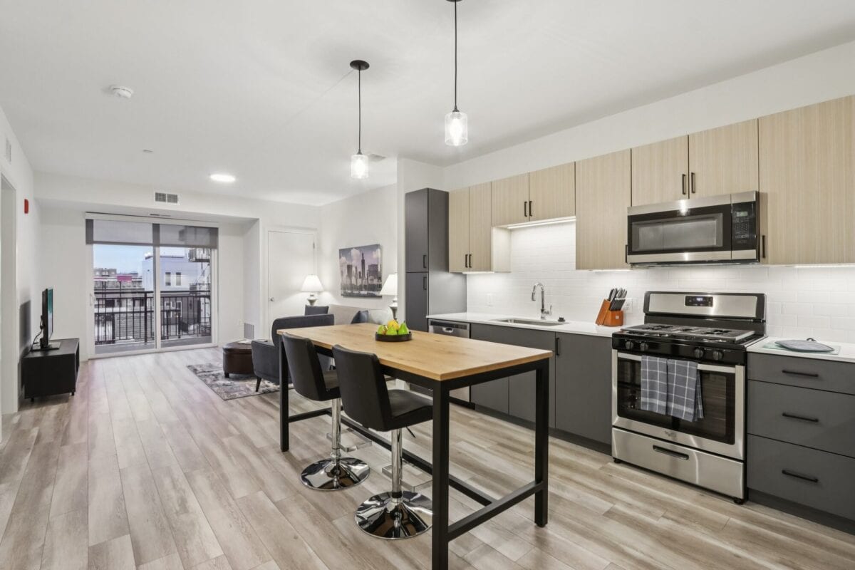 Modern apartment kitchen with light wood floors, island dining table with two chairs, stainless steel appliances, and a view into the living area with balcony access.