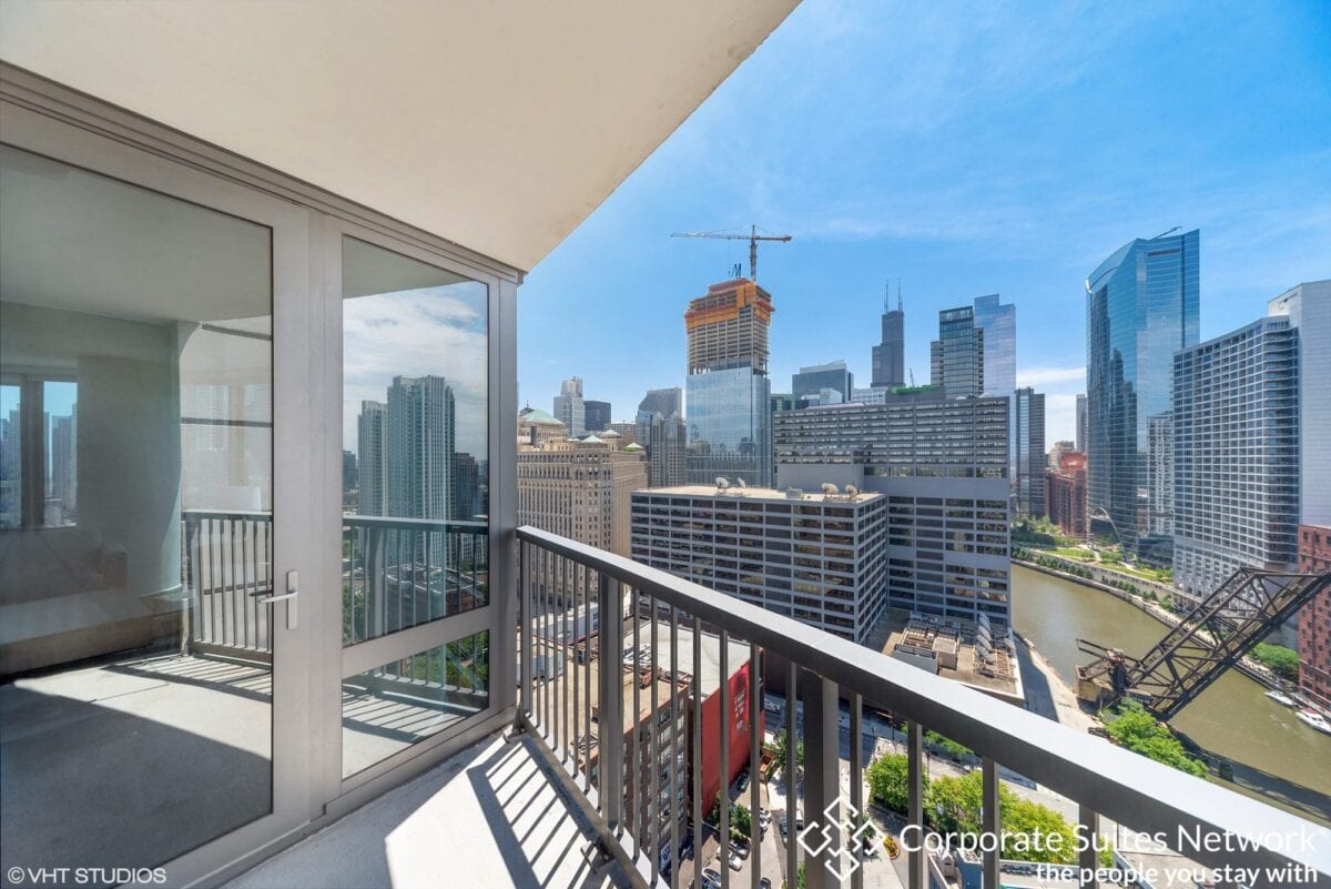 High-rise balcony view overlooking downtown city buildings, a river, and a construction crane, with clear blue sky above. Part of a glass door and the interior of an apartment are visible.