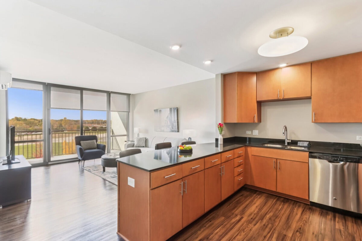 Modern apartment with open-concept kitchen and living area, wood floors, stainless steel appliances, and large windows overlooking a balcony with outdoor views.