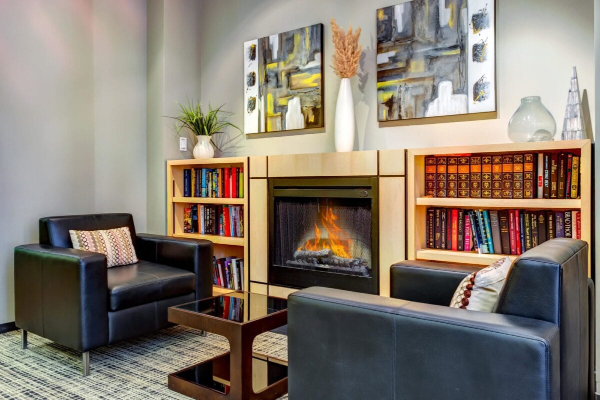 Modern living room with two black armchairs, a glass coffee table, built-in bookshelves, a fireplace, abstract art, and decorative vases on the mantel.