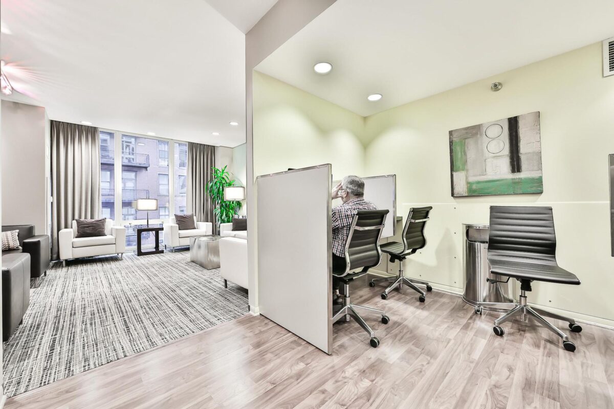 Modern office space with private cubicles, swivel chairs, and a lounge area featuring armchairs, large windows, and neutral decor.
