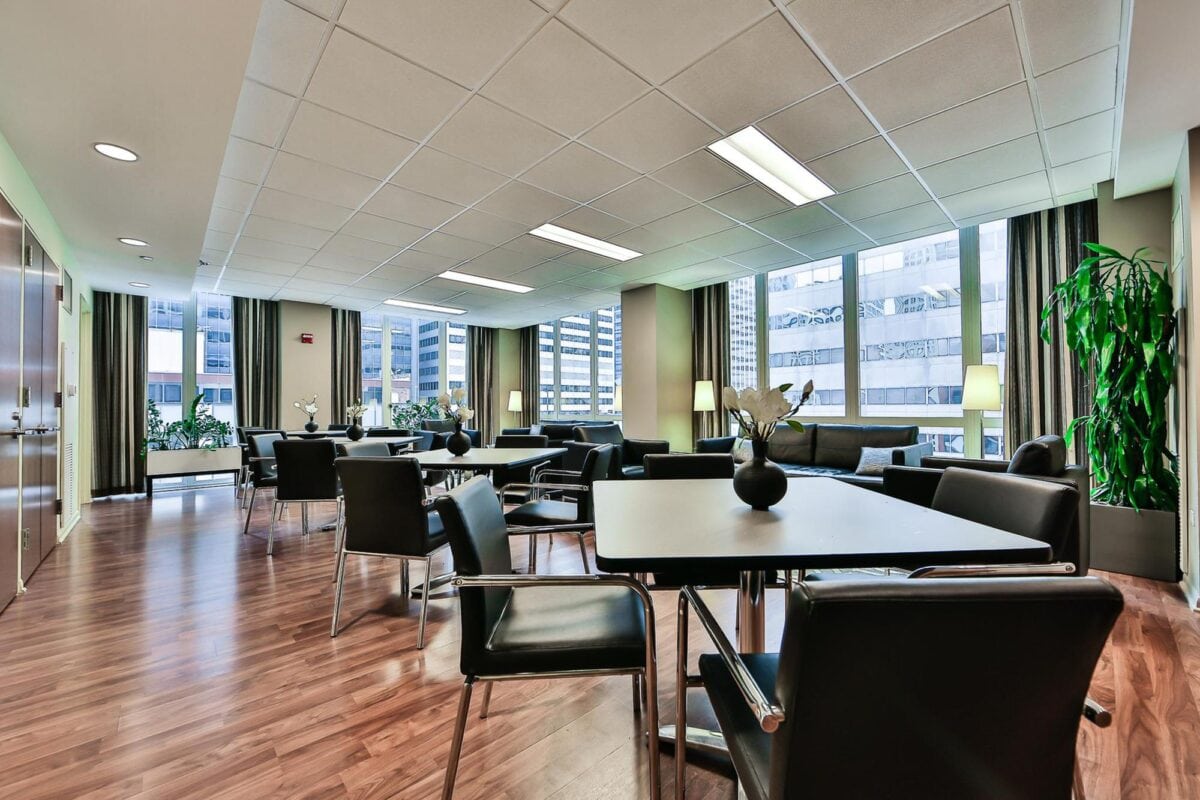 Modern office break room with several tables and chairs, large windows, potted plants, and wood flooring; city buildings visible through the windows.