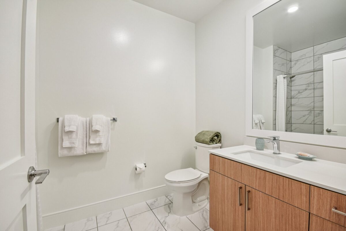 Modern bathroom with light wood vanity, white sink, toilet, wall-mounted towels, and marble tile floor; shower with glass door reflected in the mirror.