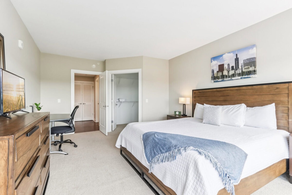 Modern bedroom with a large wooden bed, white bedding, desk and chair, dresser with TV, and a closet with double doors; wall art hangs above the bed.