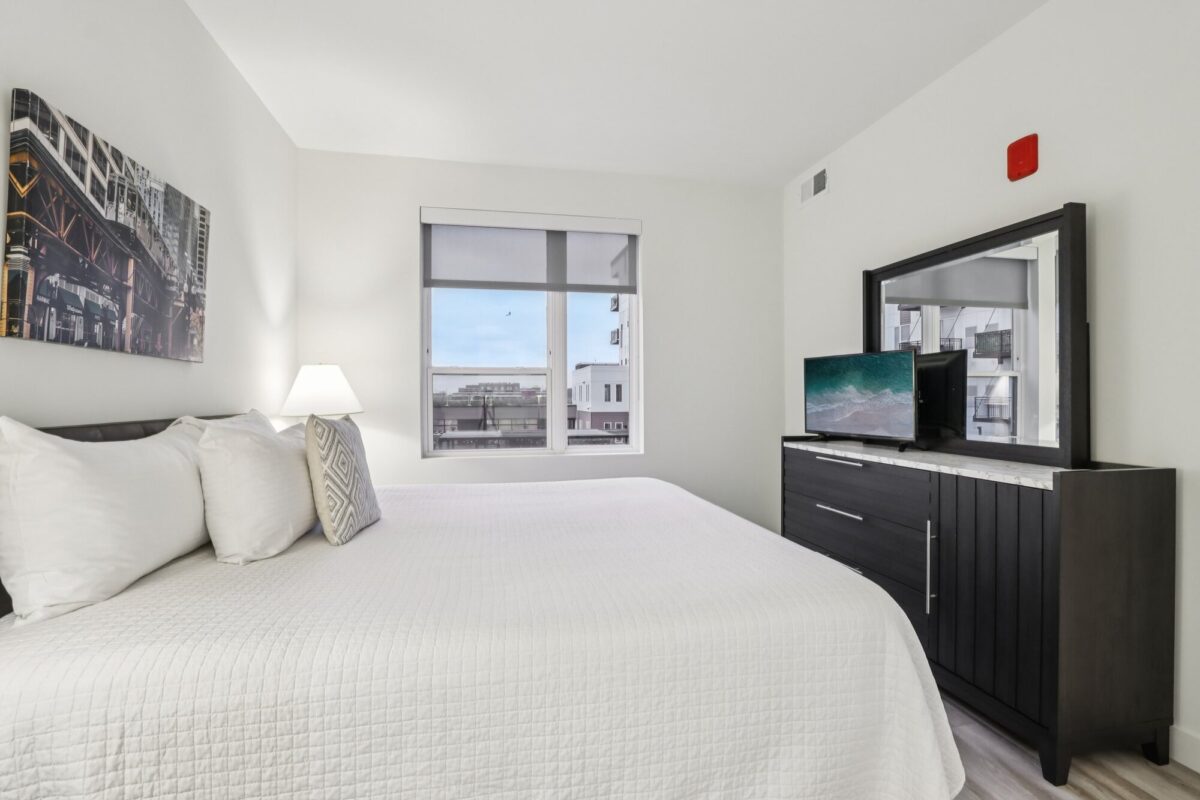 Modern bedroom with a neatly made bed, large window, dresser with a mirror, TV, and a lamp on a bedside table.