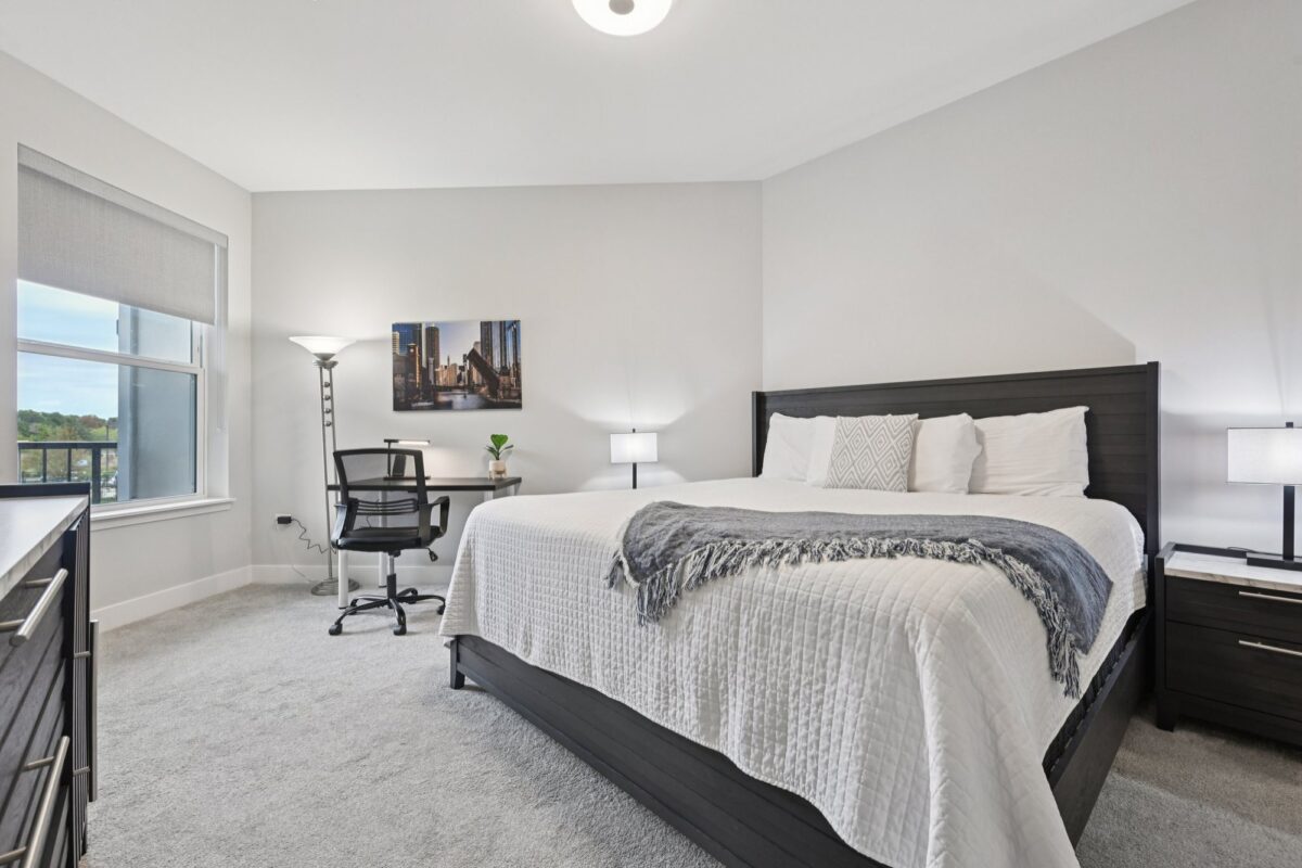 A modern bedroom with a large bed, two nightstands, a desk with a chair, a floor lamp, and a window with a city view.