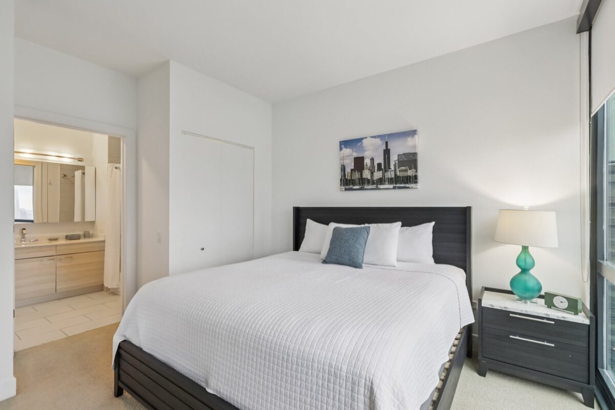 Modern bedroom with a large bed, white bedding, nightstand, lamp, cityscape artwork, and an en suite bathroom visible through an open door.