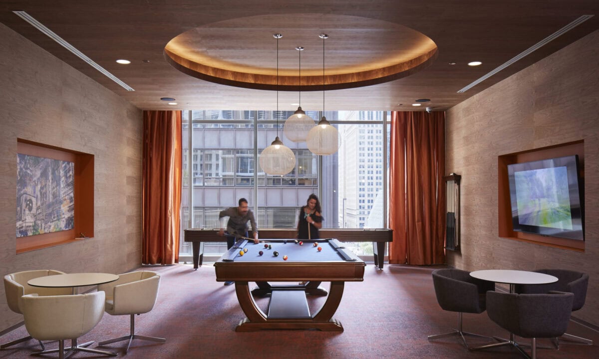 Two people play pool in a modern lounge with a large window, hanging lights, table seating, and city buildings visible outside.