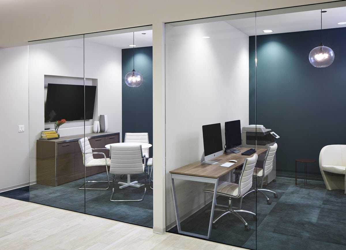Modern office interior with two glass-walled meeting rooms, each furnished with desks, chairs, computers, a TV, and pendant lights.
