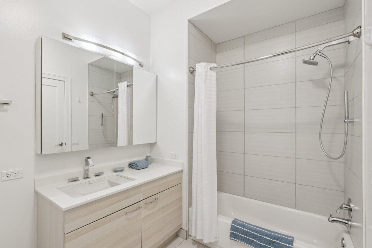 Modern bathroom with a light wood vanity, double sink, large mirror, bathtub with shower, white curtain, and a blue towel. Walls and tiles are light-colored for a clean look.