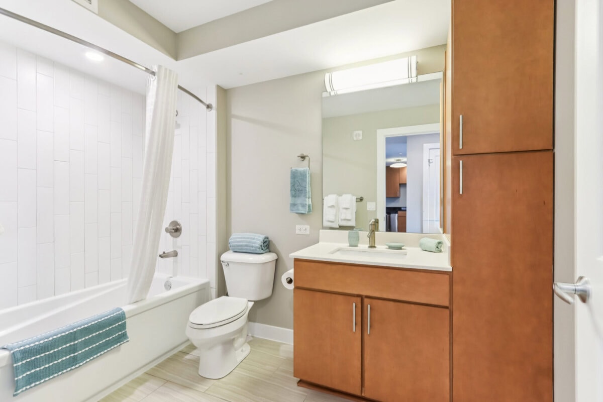 Modern bathroom with a bathtub, shower curtain, toilet, sink, large mirror, wooden cabinets, and light-colored tile flooring. Blue towels and decor accents are present.