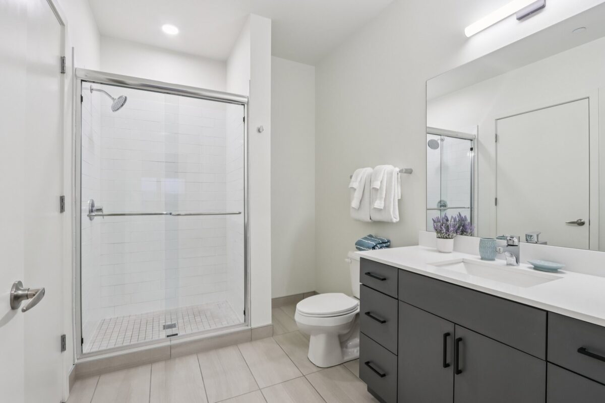 Modern bathroom with a glass shower, white toilet, dark vanity with sink and mirror, towel rack, and neutral-colored tile floor.