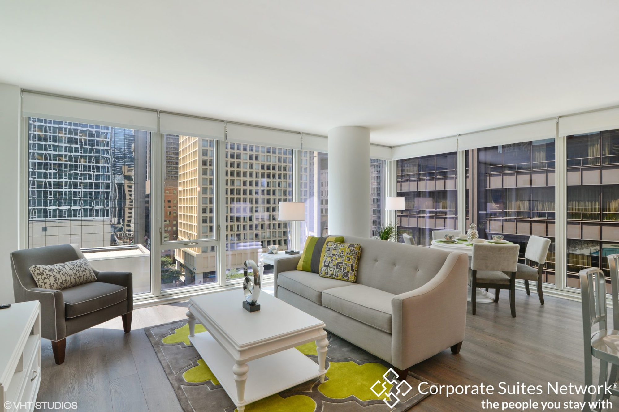 Modern high-rise apartment living room with large windows, neutral furniture, city views, and a dining area in the background. Corporate Suites Network logo is visible in the corner.
