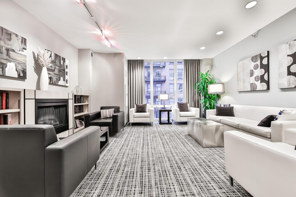 Modern living room with white and black furniture, abstract wall art, a fireplace, bookshelves, large windows with city view, potted plant, and patterned carpet.
