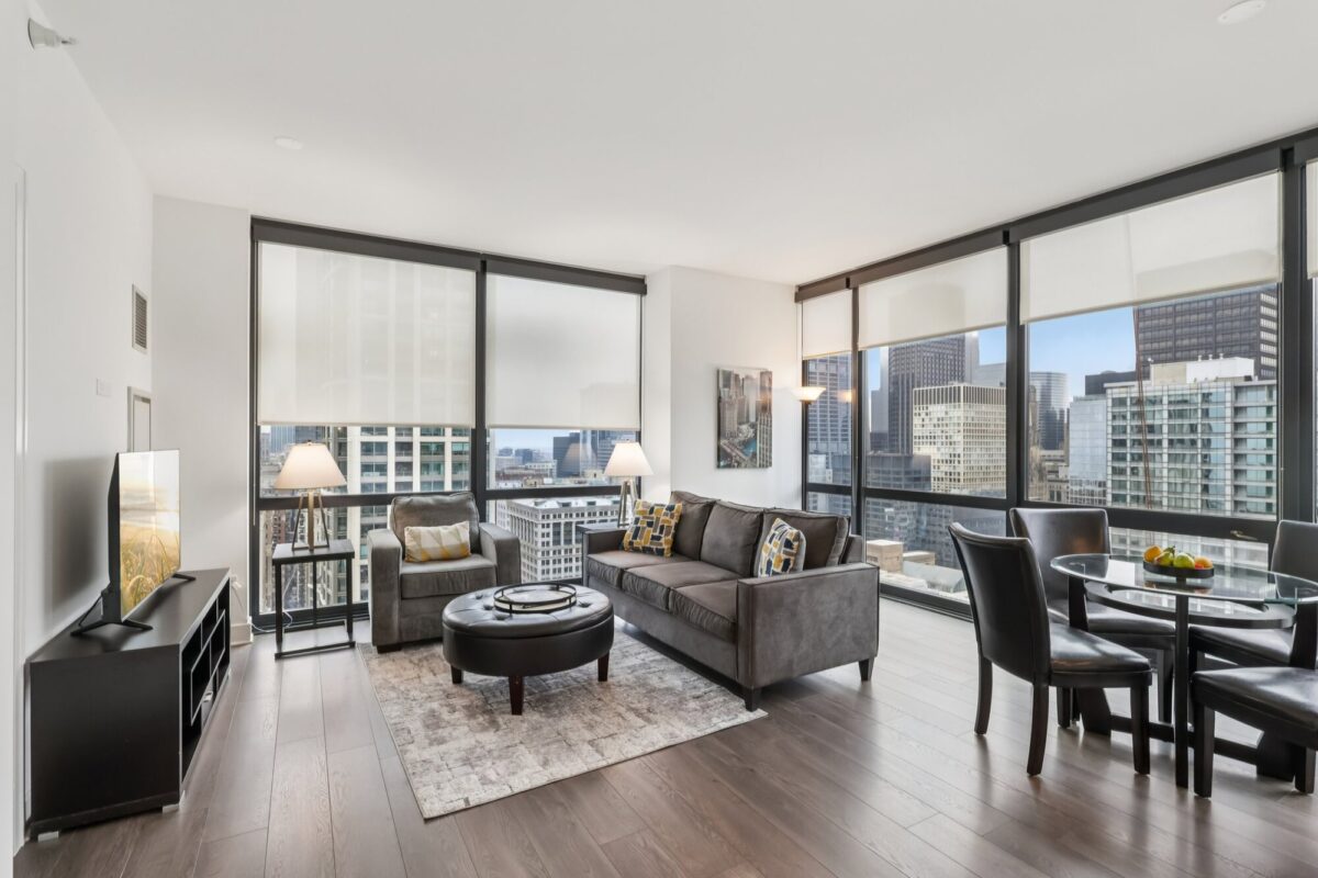 Modern high-rise apartment living room with large windows, gray sofas, a round coffee table, TV, dining area, and city skyline views.