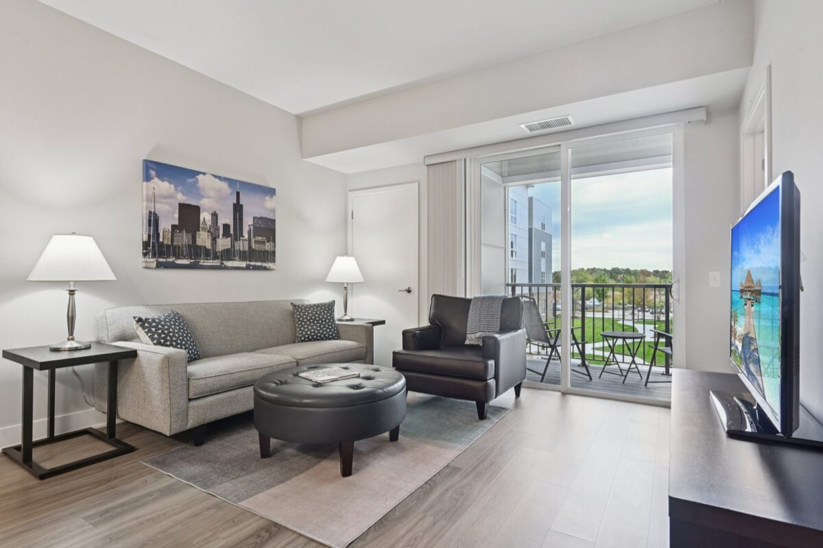 Modern living room with a sofa, armchair, round ottoman, two side tables with lamps, wall art, TV, and sliding glass doors leading to a balcony with outdoor seating.