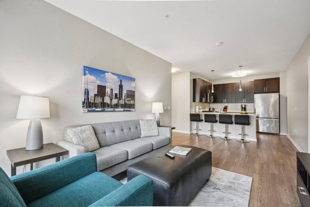 Modern open-concept living room and kitchen with gray sofa, teal armchair, wall art, bar stools, and stainless steel appliances.