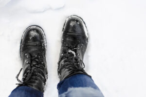 A person wearing black lace-up boots and blue jeans stands on snow-covered ground, with snow clinging to the boots.