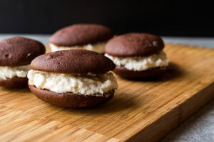 Four chocolate whoopie pies with creamy white filling are arranged on a wooden cutting board, with a dark background and a focus on the front pie.