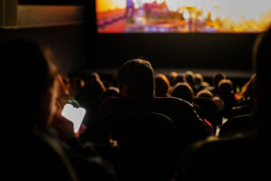 A dimly lit movie theater with people seated and watching a film; one person in the foreground holds a brightly lit phone, drawing attention in the darkness.