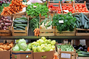 A market stall displays various fresh vegetables in wooden crates, including carrots, tomatoes, cucumbers, beets, parsley, bell peppers, cabbage, potatoes, lettuce, and eggplants.