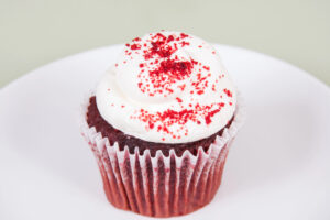 A red velvet cupcake with white frosting and red sprinkles sits on a white plate against a light background.