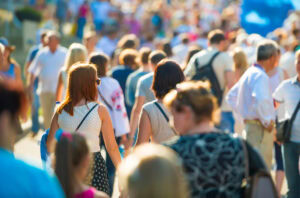 A large crowd of people walking outdoors on a sunny day, with two women in the foreground holding hands. The scene is lively and busy, with many individuals in casual summer clothing.