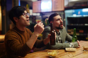 Two men sit at a bar, watching something offscreen with excited expressions. One holds a beer bottle and the other has popcorn in hand. Snacks and drinks are on the wooden counter in front of them.