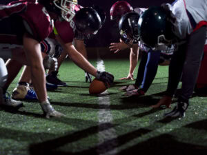 Two American football teams face each other at the line of scrimmage, crouched and ready to play, with one player holding the ball on a lit field at night.