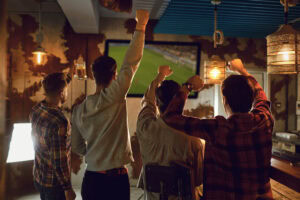 Four men stand with raised arms, cheering while watching a sports match on a large TV in a dimly lit bar with rustic decor and hanging lights.
