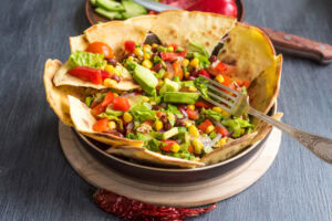 A tortilla bowl filled with a colorful salad of lettuce, avocado, cherry tomatoes, corn, kidney beans, and red onions, with a fork resting on the side and vegetables in the background.