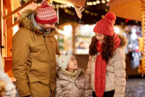 A family of three, dressed in warm winter clothes and hats, smiles and looks at each other while standing outdoors at a festive holiday market with lights in the background.