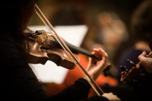 Close-up of a person playing a violin, focusing on their hand pressing the strings and the instrument’s body. The background is blurred, highlighting the musician and the violin.