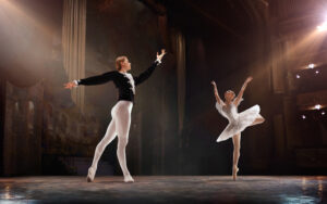 Two ballet dancers perform on stage; the male dancer wears a black costume and stands en pointe with one arm raised, while the female dancer in a white tutu poses gracefully on one leg, lit by stage lights.