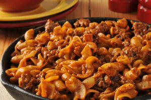 A close-up of a skillet filled with beef and pasta noodles in a rich, red-brown sauce, sitting on a wooden table. The dish looks hearty and flavorful.