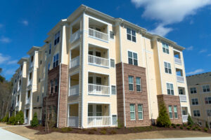 Four-story modern apartment building with light yellow siding, white trim, and brick accents. Each unit has a balcony, and the landscaping includes small bushes and neatly trimmed grass under a partly cloudy sky.