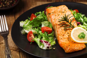 Grilled salmon fillet served on a bed of fresh mixed salad greens with cherry tomatoes, garnished with a lemon slice and rosemary sprig, on a black plate.