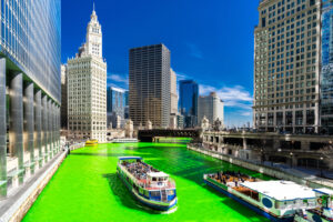 A vibrant green river runs through a city with tall buildings, boats carrying people, and a clear blue sky overhead. The scene appears festive and lively.