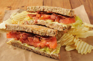 A BLT sandwich with bacon, lettuce, and tomato on whole grain bread, cut in half and served on brown paper with a side of crinkle-cut potato chips.